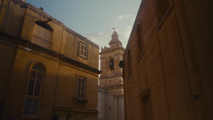 A low-angle shot captures a historic church bell tower framed by ancient, sun-drenched stone buildings