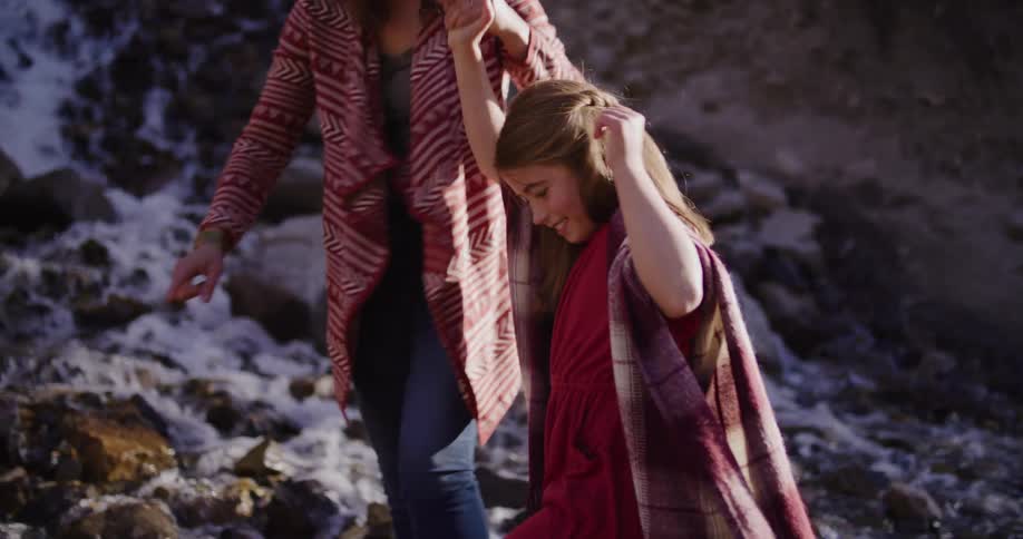 A caring mother holds her smiling daughter's hand, guiding her across a rocky stream in slow motion