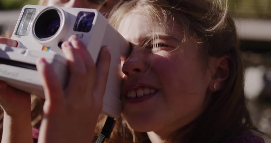A smiling young girl uses an instant camera to take a picture, guided by her happy mother in a heartwarming slow-motion close-up