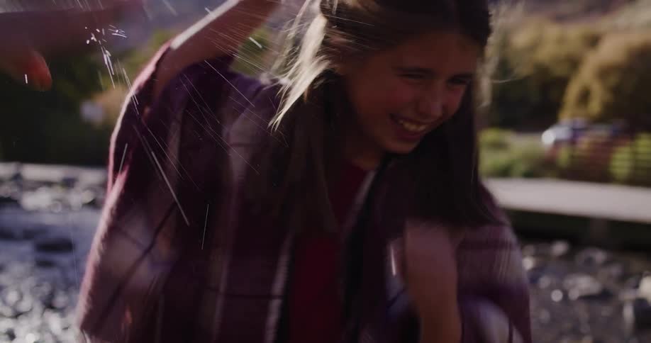 Smiling girl in a plaid shawl splashes water while playing in a shallow stream in slow motion
