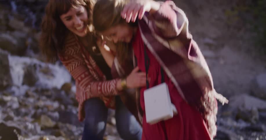 A happy mother helps her laughing daughter as they carefully cross a rocky stream together in nature