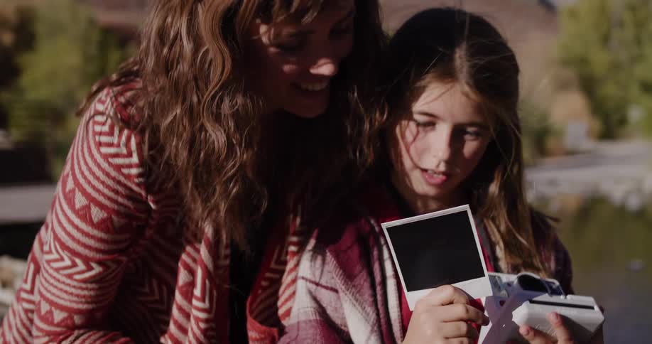 A young girl eagerly shakes an instant photo as it develops, sharing the moment with her smiling mother outdoors