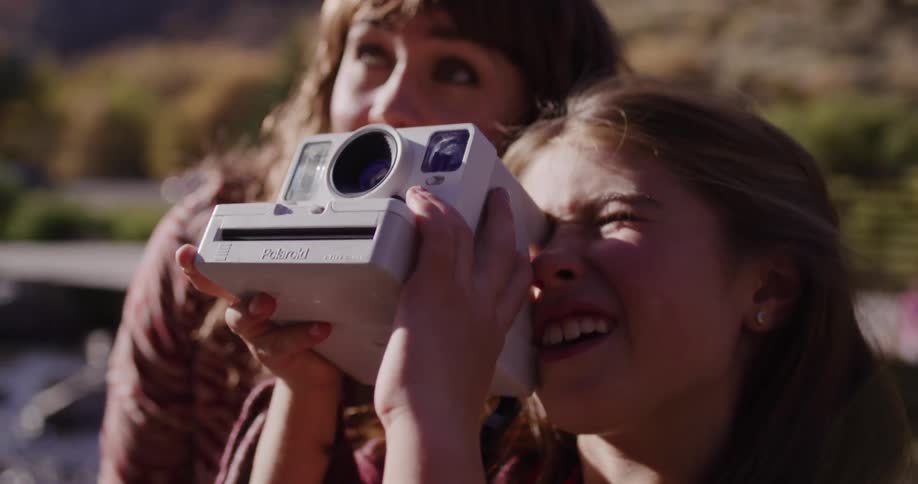 A mother helps her smiling daughter take a picture with a Polaroid instant camera outdoors