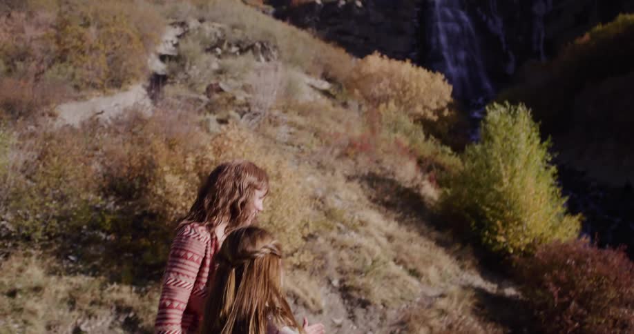 A mother shows her daughter a stunning waterfall while hiking together through colorful autumn mountains