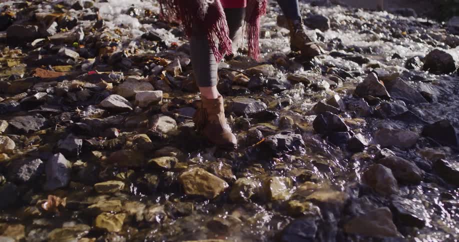 A woman in fringed boots carefully steps across a shallow, rocky stream in slow motion on a sunny day