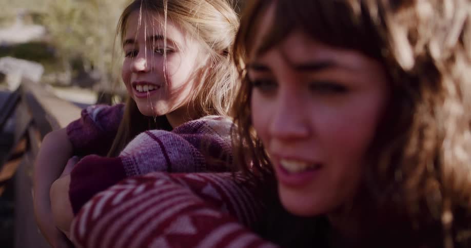 A young woman and a girl lean on a wooden railing outdoors, sharing a quiet, smiling moment in the sun