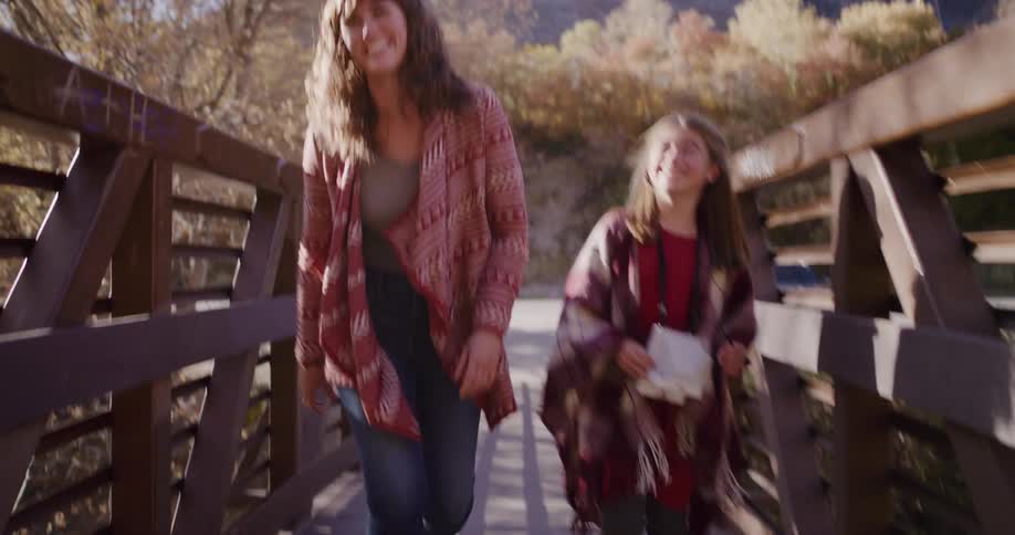 A smiling mother and her young daughter walk together across a wooden bridge on a sunny day in the mountains