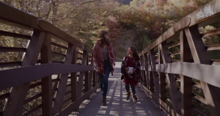 A smiling mother and daughter walk together across a wooden bridge surrounded by colorful autumn trees