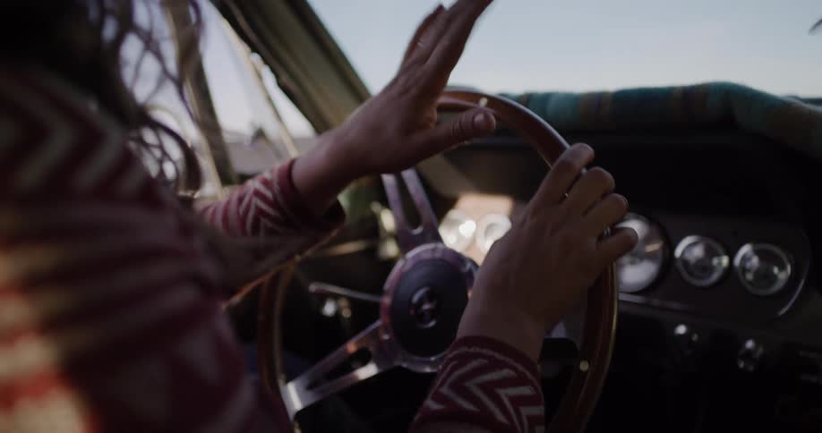 A woman taps her hands to the music on the steering wheel of a classic car during a sunny road trip