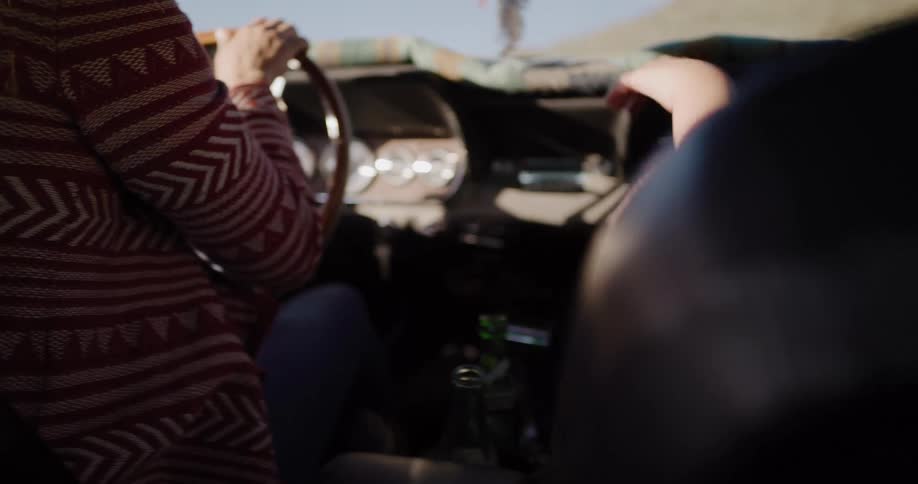 Two women in bohemian-style clothing dance and vibe to the music from the front seat of a vintage convertible during a sunny road trip
