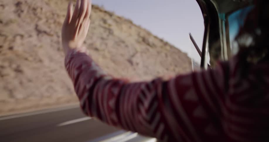 A woman holds her hand out the window of a moving car, feeling the wind as she drives along a desert highway in slow motion