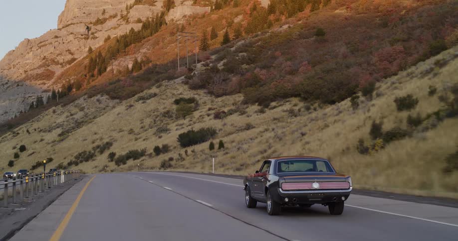 A vintage black muscle car cruises along a winding mountain highway as the sun sets