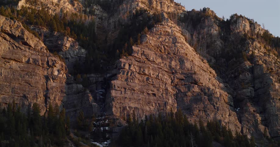 Golden hour light illuminates the rugged cliffs and forests of a massive mountain in a slow aerial tilt
