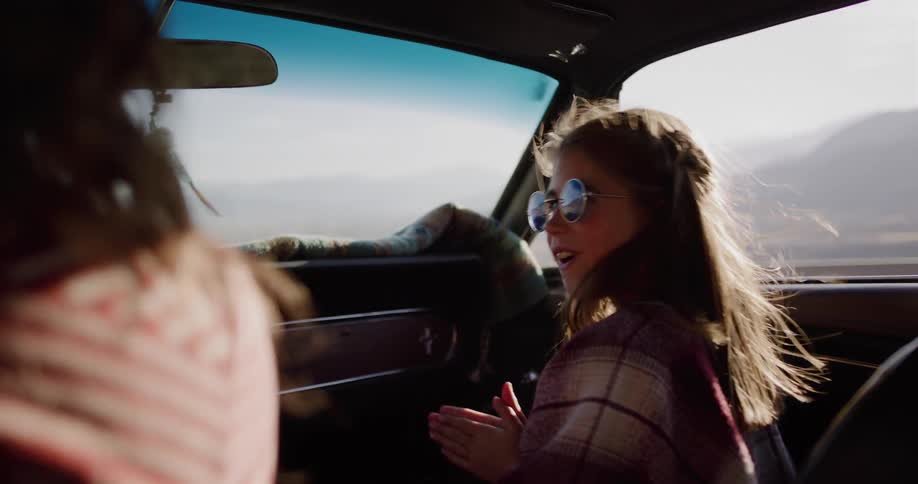 Shot from the backseat of a vintage car, a happy young girl in sunglasses claps and smiles during a sunny road trip