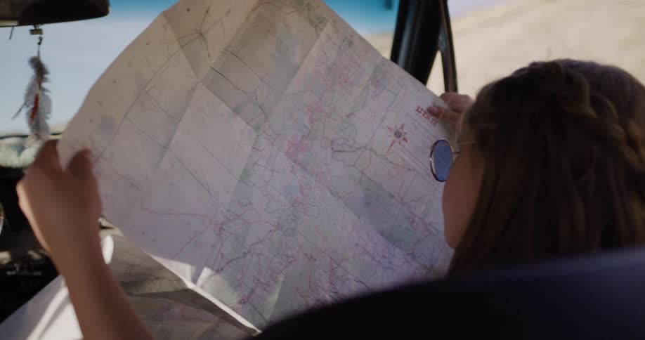 Over-the-shoulder view of an adventurous young woman studying a large paper map of Utah from the passenger seat of a car