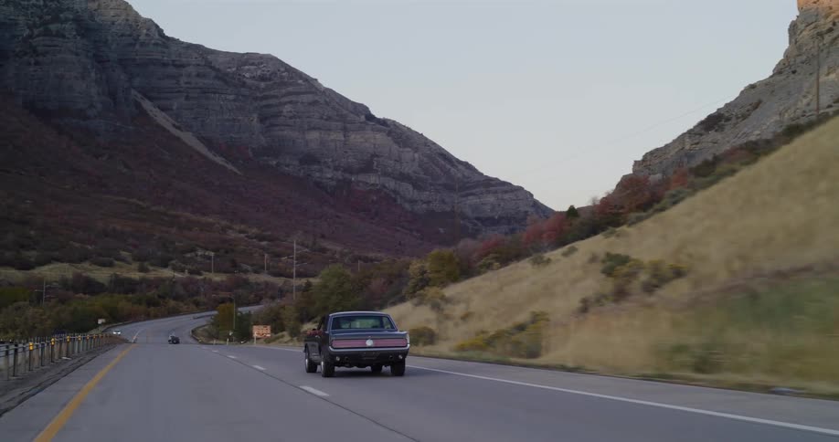 A classic muscle car cruises down a scenic mountain highway during a beautiful twilight evening