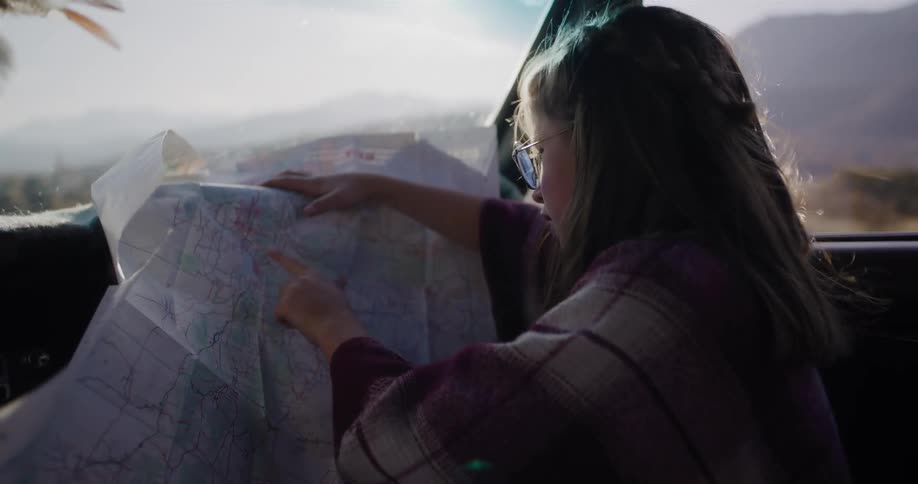 A young woman navigates with a paper map from the passenger seat of a vintage car during a sunny road trip through the mountains