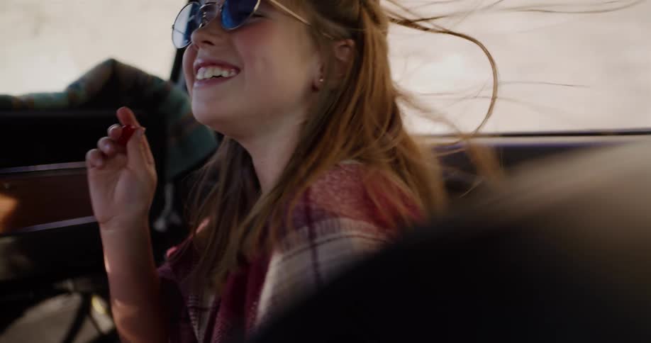 A joyful young girl wearing retro sunglasses smiles as the wind blows through her hair in a classic convertible