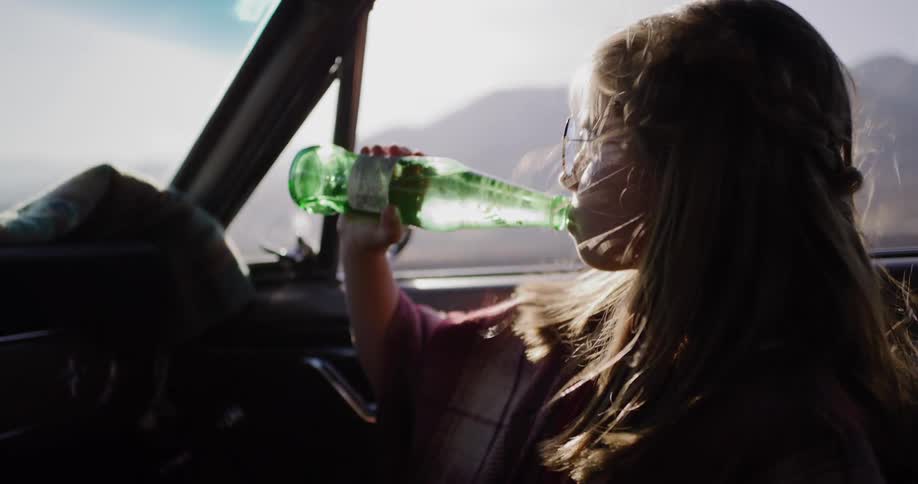 Slow motion close up of a young girl in sunglasses drinking from a green glass soda bottle during a sunny road trip