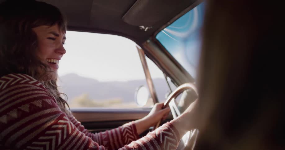 A young woman screams and laughs with uninhibited joy while driving a vintage car on a sunny road trip in slow motion