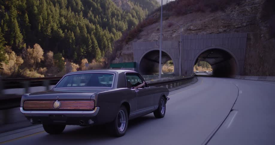A classic muscle car drives along a winding mountain highway before entering a tunnel