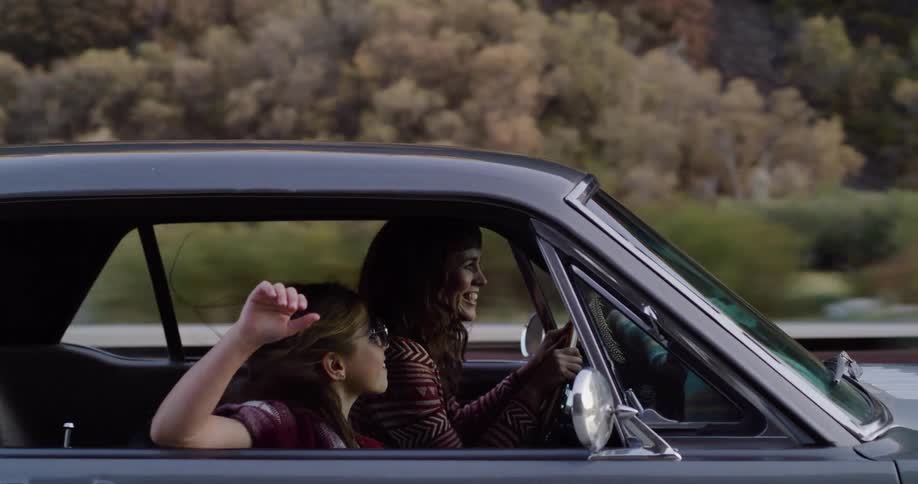 A happy young girl with sunglasses waves her hand out the window of a classic car while her mother drives them on a scenic road trip in slow motion
