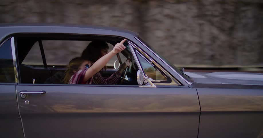 A young girl with sunglasses happily leans out the passenger window of a vintage car as her mother drives them along a scenic road