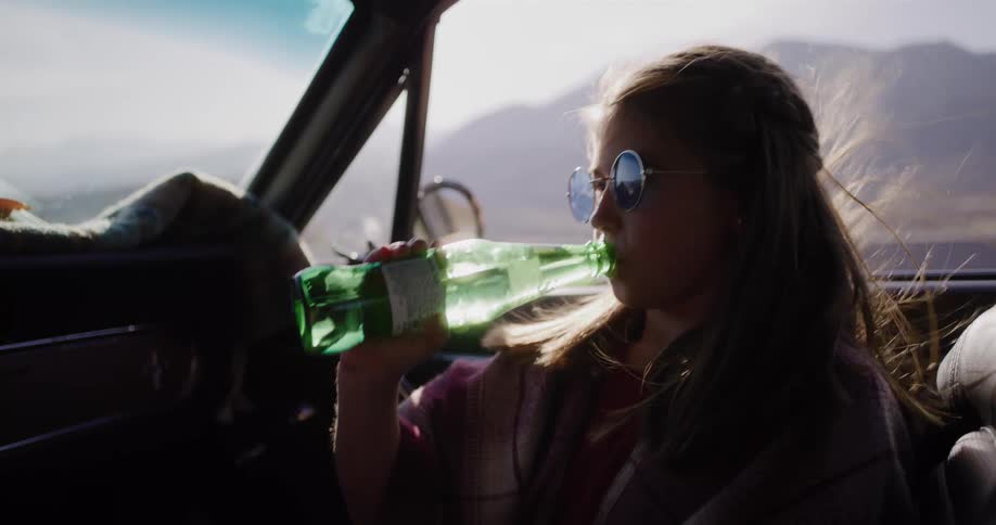 A cool girl in retro sunglasses drinks from a glass bottle in slow motion while on a scenic road trip