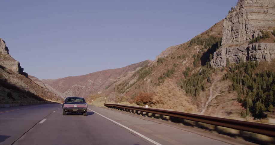 A vintage purple muscle car cruises down a scenic highway through a mountain canyon on a sunny day