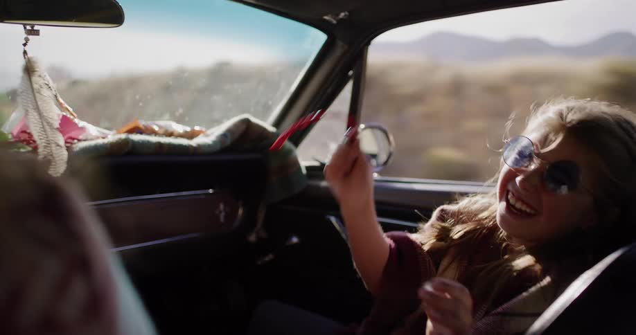 A carefree young girl wearing round sunglasses joyfully plays with a red ribbon while on a sunny road trip in a vintage car