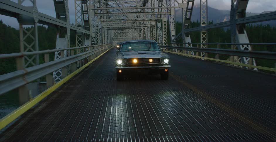 A classic Ford Mustang with headlights on drives across a steel truss bridge surrounded by a mountain forest at twilight