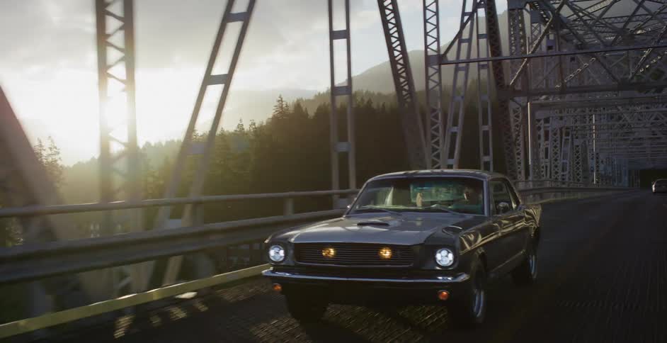 A couple enjoys a scenic drive in a classic muscle car across a steel truss bridge as the sun rises over the mountains