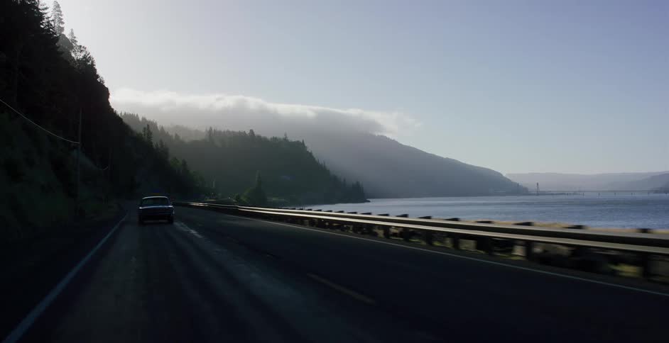 A scenic drive on a highway alongside a wide river with foggy mountains in the background during sunrise