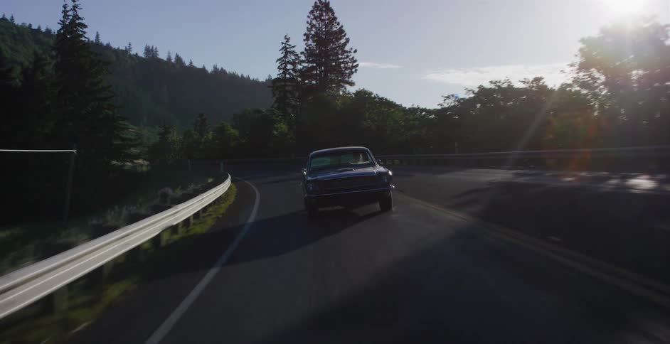 Front view of a classic black Mustang driving on a winding forested road at sunrise