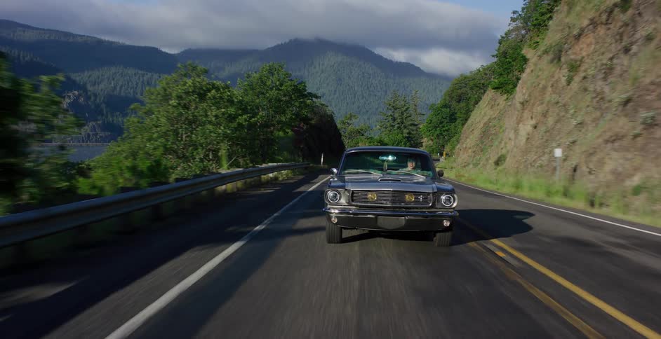 A classic Ford Mustang drives along a scenic highway with mountains and a river in the background