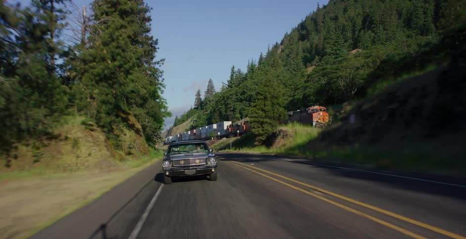 A vintage muscle car drives down a scenic mountain highway as a long freight train travels along a parallel track on a sunny day