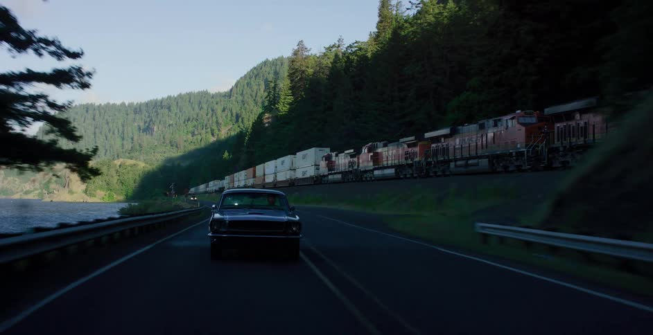 A vintage black muscle car drives along a scenic highway beside a moving freight train before entering and emerging from a dark tunnel