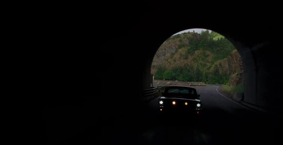 A classic Ford Mustang drives along a scenic highway next to a river before exiting a mountain tunnel in a beautiful natural landscape