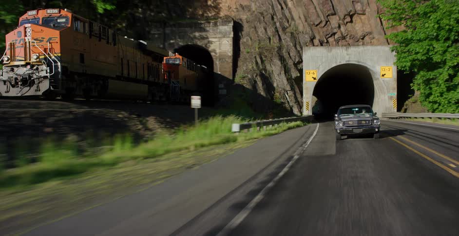 A tracking shot pulls back from a dark tunnel to reveal a vintage car driving on a mountain road as a freight train emerges from a parallel tunnel