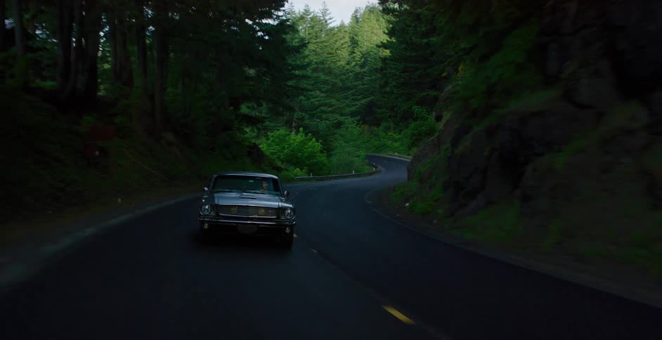 A vintage muscle car drives along a winding, tree-lined road through a dense, dark forest