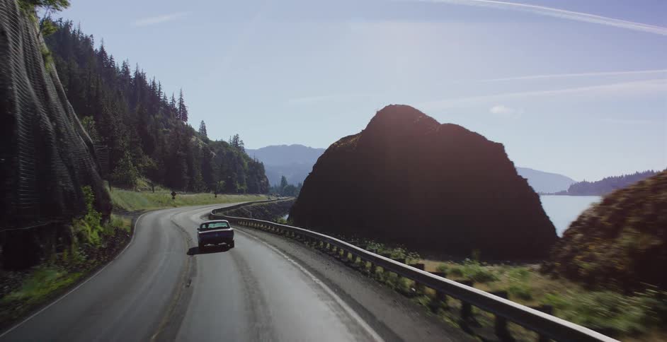 A slow-motion shot follows a vintage car as it emerges from a dark tunnel onto a beautiful riverside highway on a sunny day