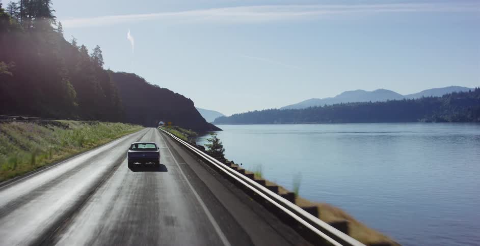 A classic dark blue convertible car drives down a scenic highway alongside a wide, calm river on a beautiful sunny day
