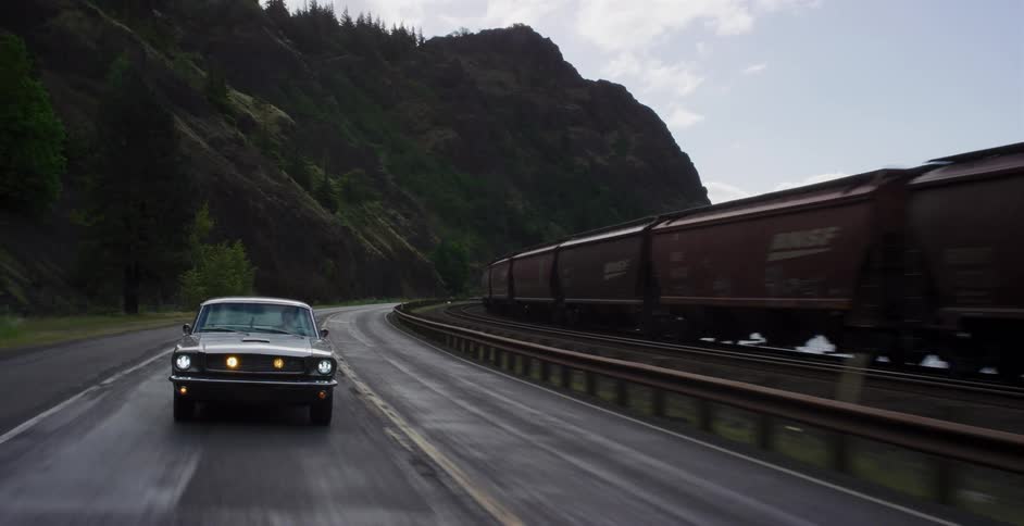 A classic Ford Mustang drives along a wet, winding mountain highway parallel to a moving freight train