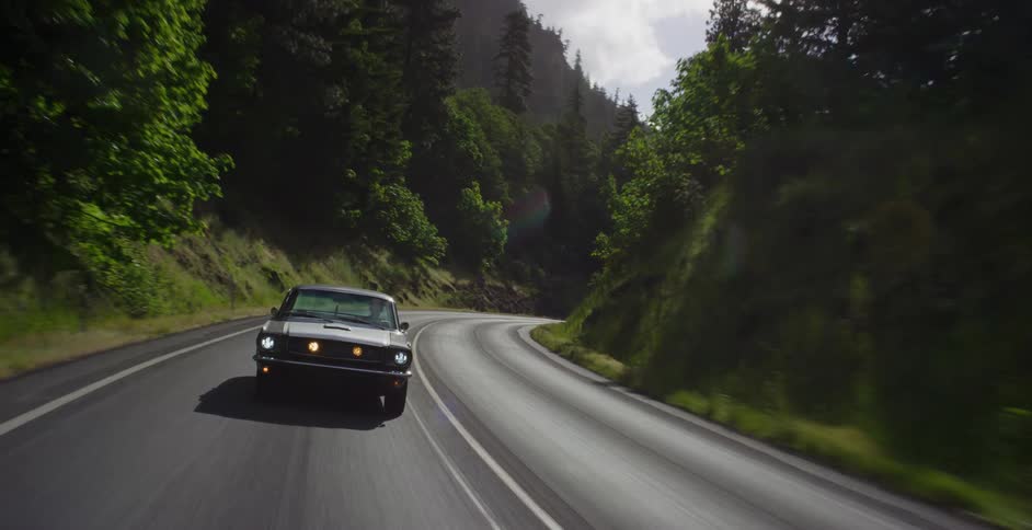 A vintage muscle car cruises along a winding scenic highway surrounded by a lush green forest on a sunny day