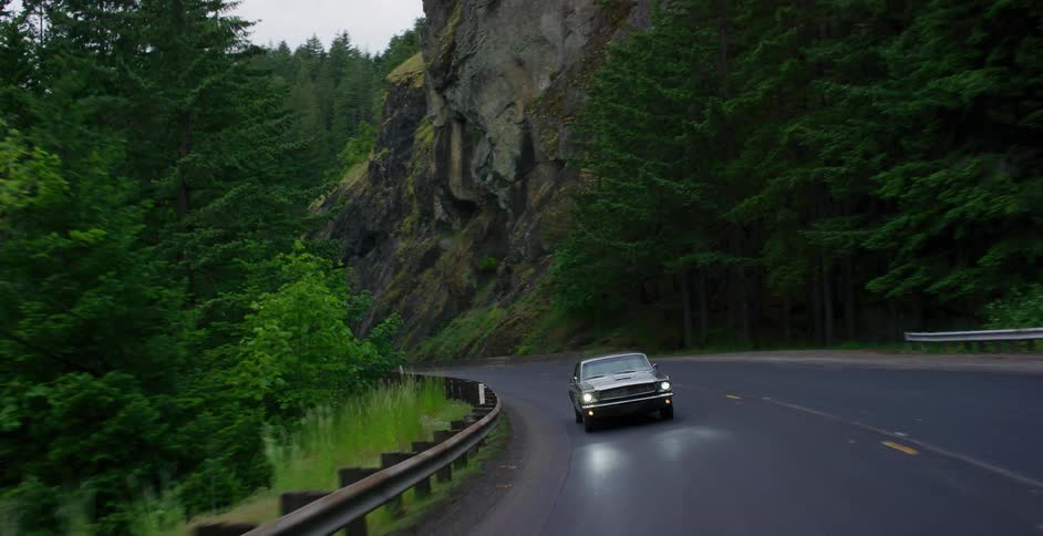 A classic muscle car with headlights on drives down a winding mountain road through a dense green forest