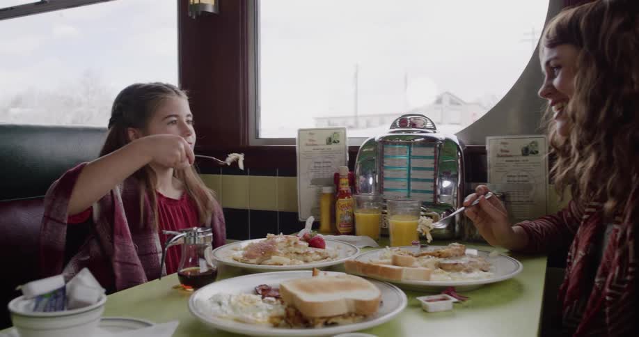 A cute young girl smiles as she eats breakfast with her mother in a cozy diner booth
