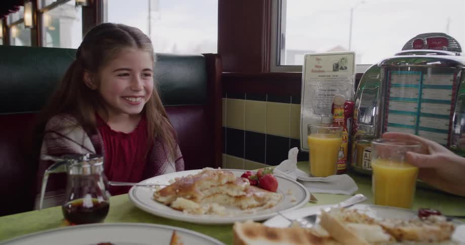 An adorable young girl expresses pure joy and excitement as she sits down for a pancake breakfast in a classic diner