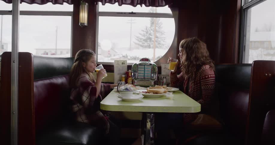 A mother and daughter share a toast with orange juice while having breakfast in a cozy retro diner on a snowy day
