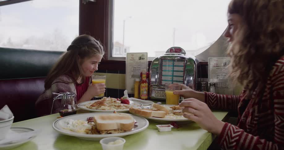A happy young girl and her mother share a smile while drinking orange juice and eating breakfast at a classic diner