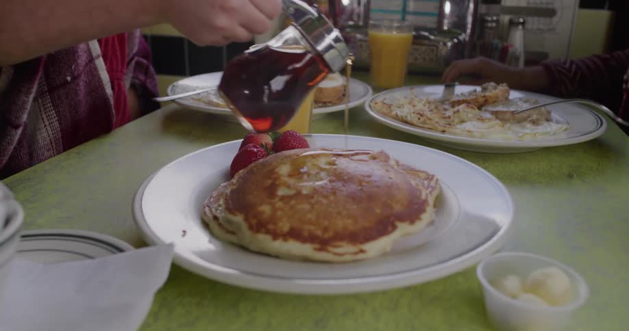A young girl pours maple syrup over a large stack of pancakes for breakfast at a retro American diner